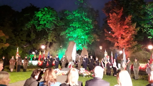 Delegacja strażaków mazowieckich przy pomniku upamiętniającym powstańców Mokotowa, w tle zielone i czerwone światło.
