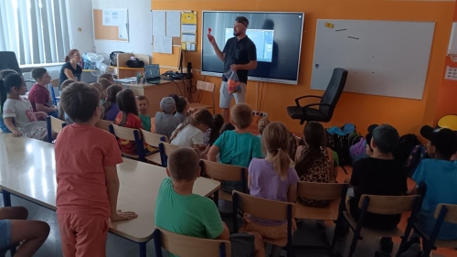 Uczniowie siedzą przed tablicą multimedialną, nauczyciel siedzi przy biurku. Gość pokazuje rzeczy, jakie można drukować 3D.