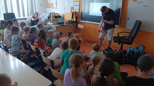 Uczniowie siedzą przed tablicą multimedialną, nauczyciel siedzi przy biurku. Gość pokazuje rzeczy, jakie można drukować 3D.