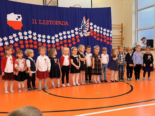 Grupa dzieci stoi na tle biało-czerwonej dekoracji. 